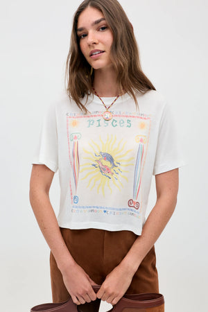 A woman with long brown hair wears the "Ria - Pisces" white T-shirt, featuring a vibrant sun and fish illustration; she pairs it with brown pants and a necklace, standing against a plain light background.