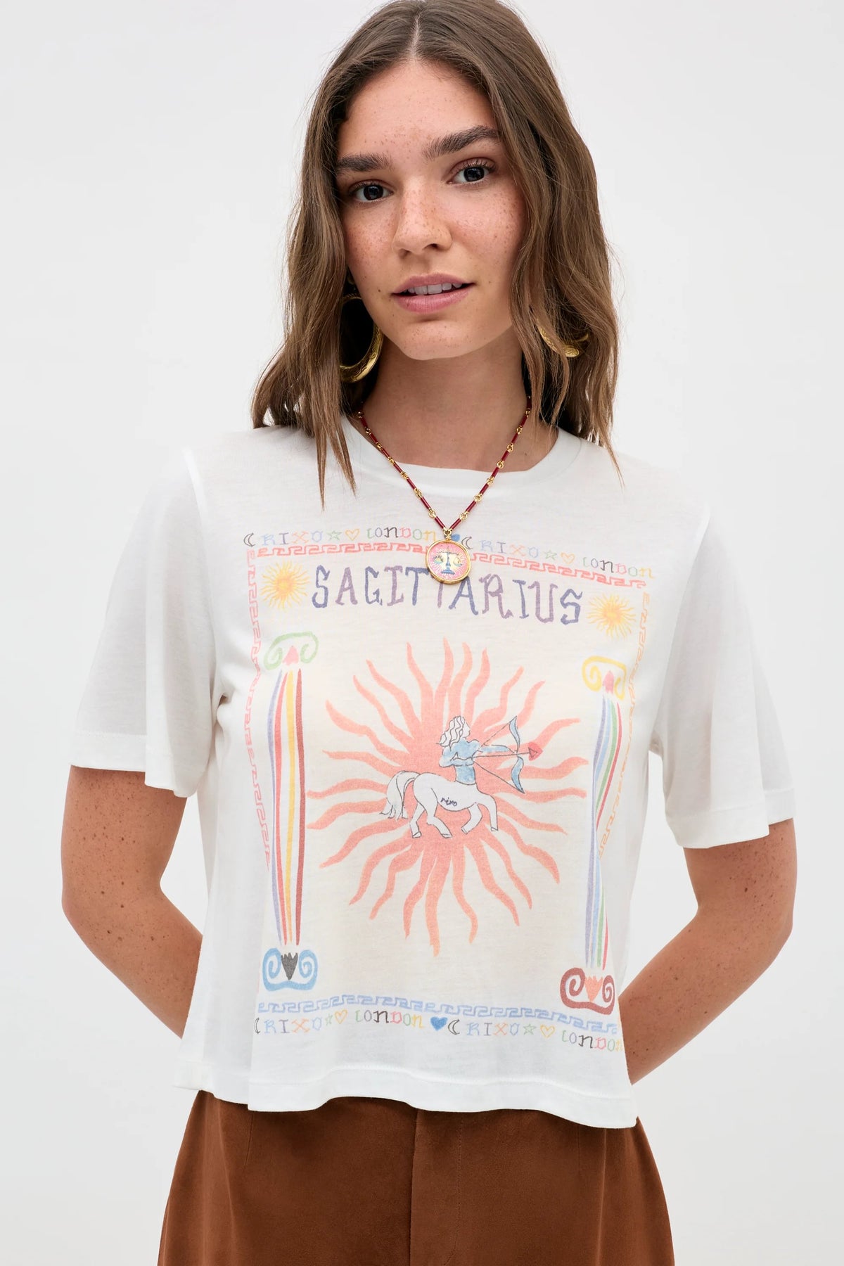 A woman with wavy brown hair models the Ria - Sagittarius white t-shirt, featuring a vibrant centaur and sun design, paired with a beaded necklace and brown pants, set against a simple background.