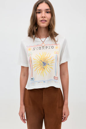 A woman with light brown hair wears the Ria - Scorpio white sheer jersey T-shirt, featuring a sun and scorpion illustration, paired with brown pants and a cropped hem, standing against a plain light background.