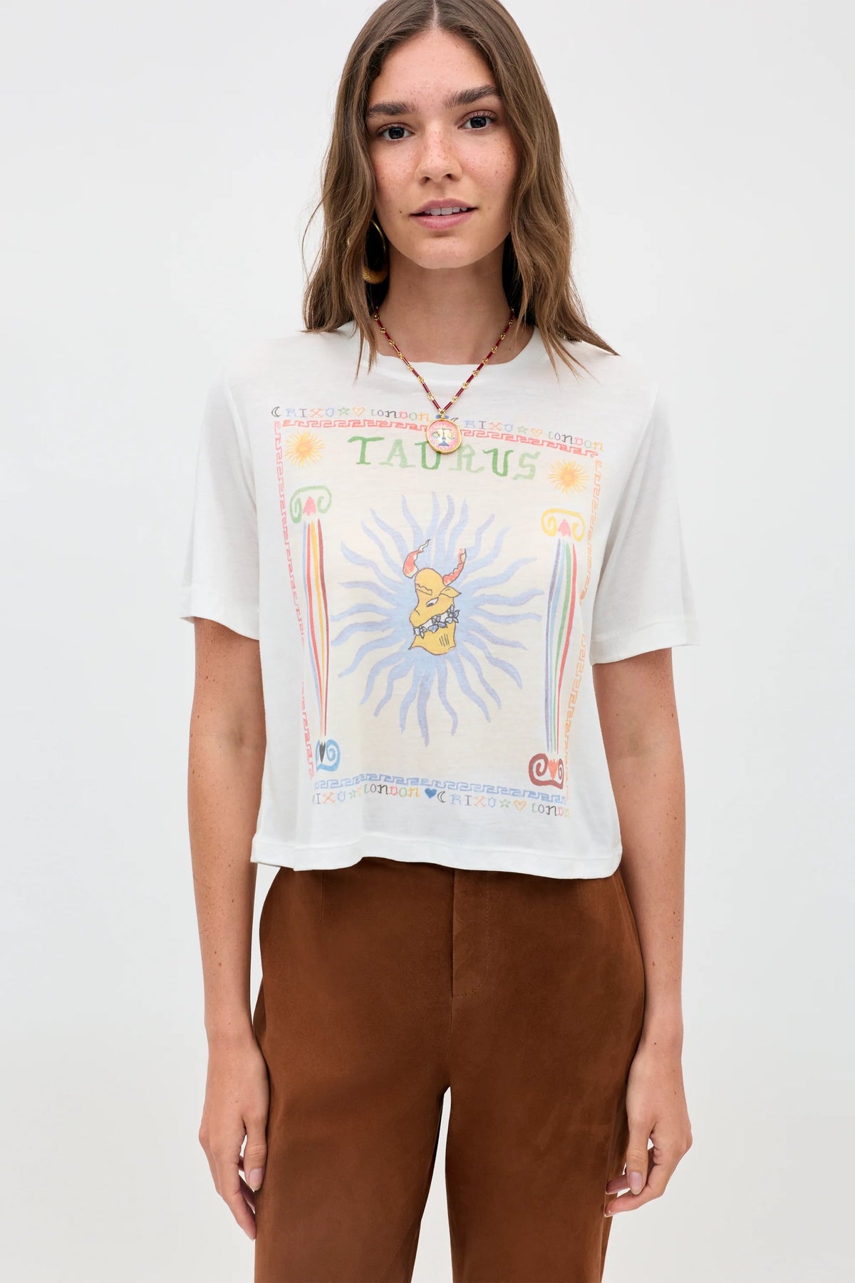 A woman with light brown hair wears the Ria - Taurus white T-shirt with a colorful Taurus design and cropped brown pants, standing against a plain light background.