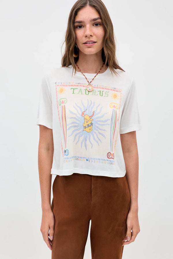 A woman with light brown hair wears the Ria - Taurus white T-shirt with a colorful Taurus design and cropped brown pants, standing against a plain light background.