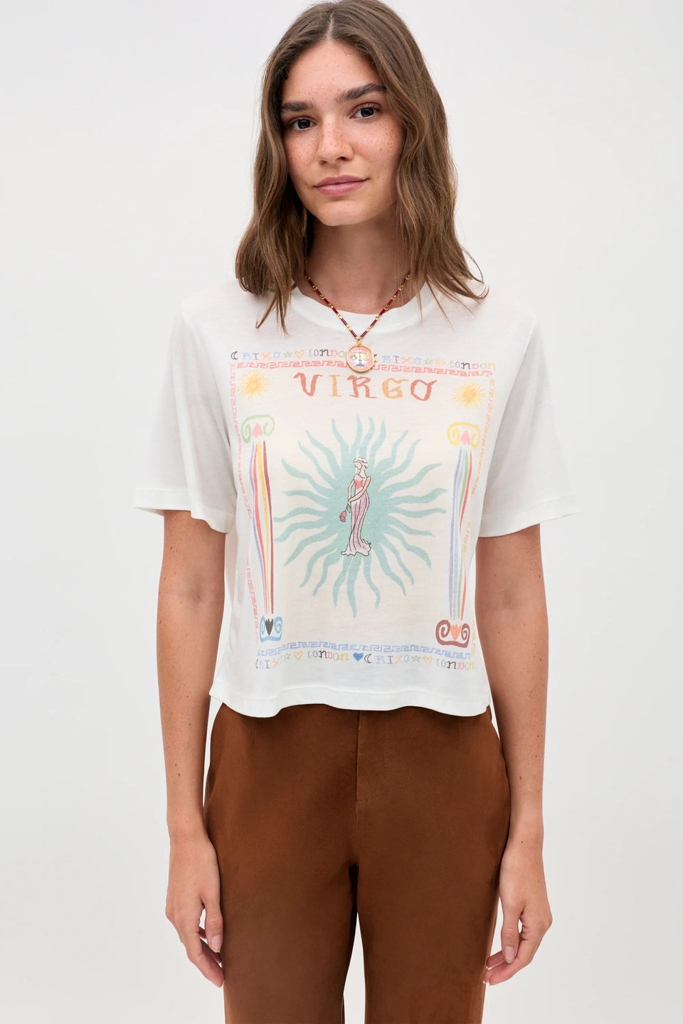 A woman with long brown hair stands against a plain light background, wearing the Ria - Virgo white T-shirt adorned with a colorful Virgo illustration and cropped-hem brown pants.