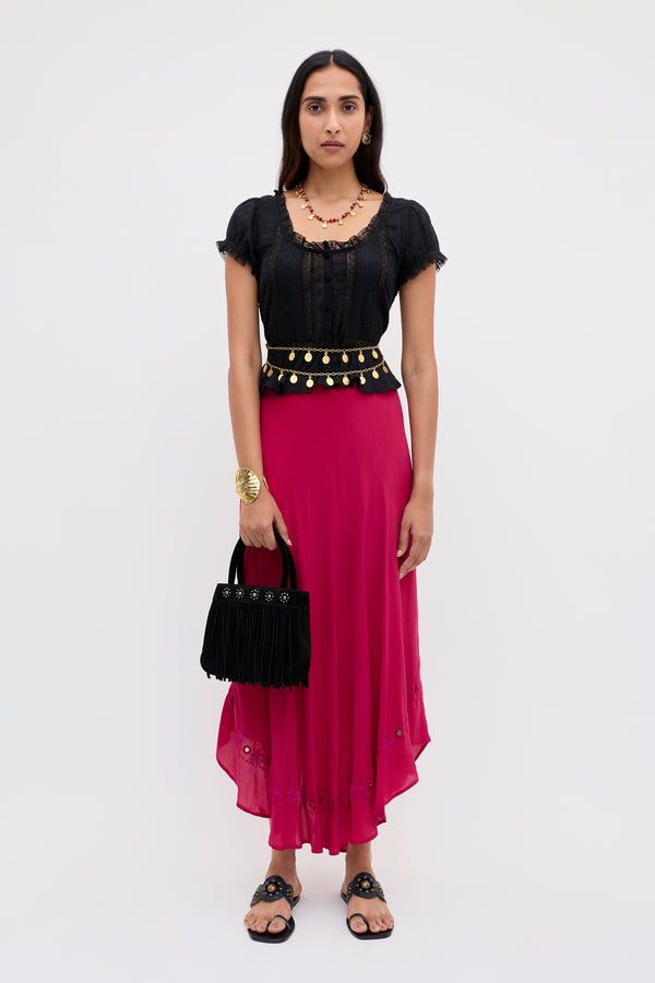 A woman stands facing forward against a plain light background, wearing the Rudy - Mirror Embellishment Magenta Pink skirt. She pairs it with a black top, black sandals, jewelry, and holds a black fringed handbag.