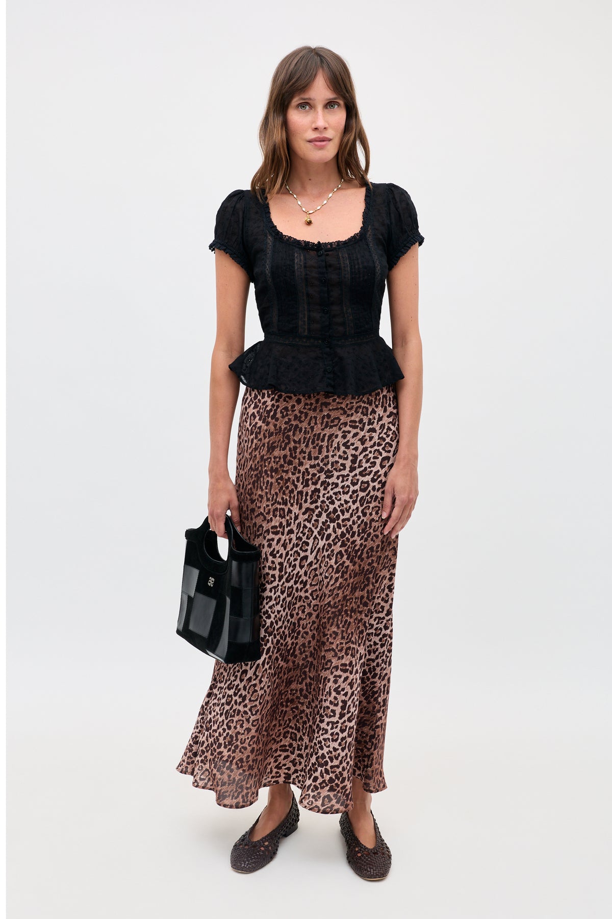 A woman with straight brown hair and a neutral expression wears the Sandi - Black short-sleeve peplum top, a long leopard print skirt, brown woven shoes, and carries a small black handbag against a plain background.