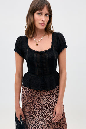 A woman with brown hair models the Sandi - Black short-sleeved blouse with lace inserts, styled with a leopard print skirt, necklace, and black bag against a light background—an ideal everyday look.