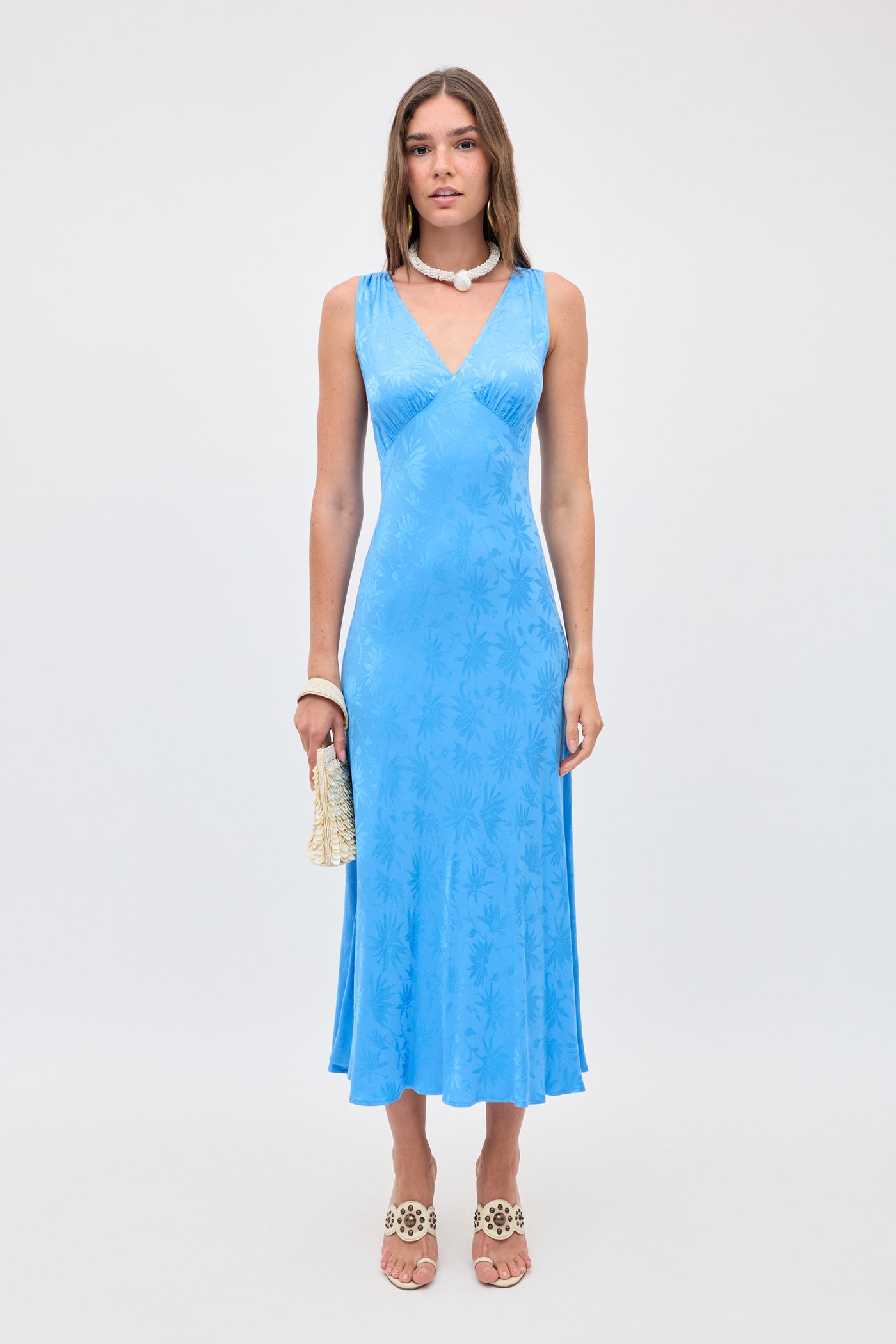 A woman stands against a plain background wearing Sandrine - Daisy Jacquard Blue, a sleeveless V-neck sky-blue slip dress with a subtle floral pattern, paired with white sandals, a pearl necklace, bracelet, and a small textured clutch.