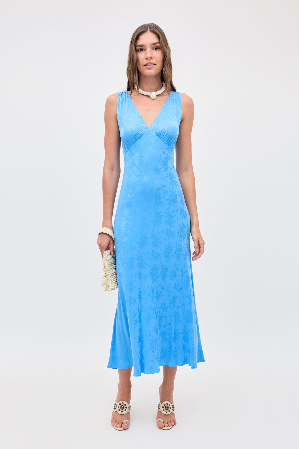 A woman in the Sandrine - Daisy Jacquard Blue slip dress, styled with white sandals, a beaded choker and bracelet, holds a woven clutch while standing against a plain background.