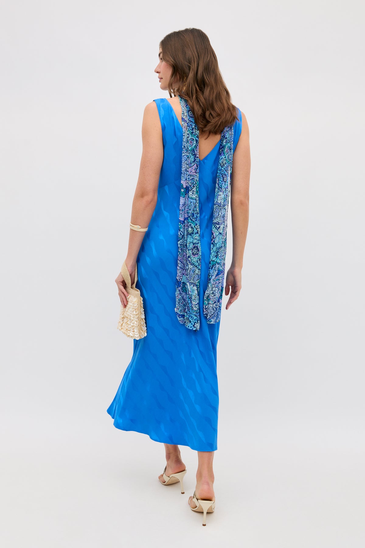 A woman in a sleeveless, bright blue Wave Jacquard Sandrine dress by RIXO pairs it with heels and a small beige handbag. A blue-patterned scarf drapes over her shoulders. Her brown hair is loose, creating a chic transitional look.