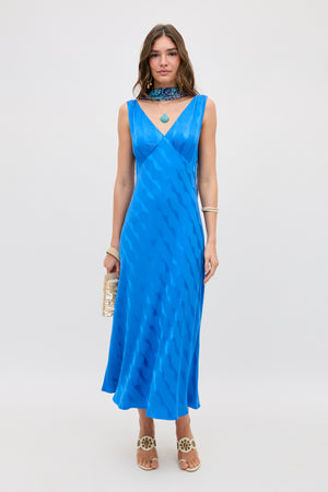 A woman wears the Sandrine - Wave Jacquard Blue dress, a sleeveless V-neck midi in bright blue with a wavy pattern, standing against a plain backdrop. She accessorizes with a gold clutch, heeled sandals, jewelry, and a choker necklace.
