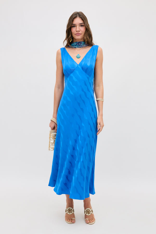 A woman wears the Sandrine - Wave Jacquard Blue dress, a sleeveless V-neck midi in bright blue with a wavy pattern, standing against a plain backdrop. She accessorizes with a gold clutch, heeled sandals, jewelry, and a choker necklace.