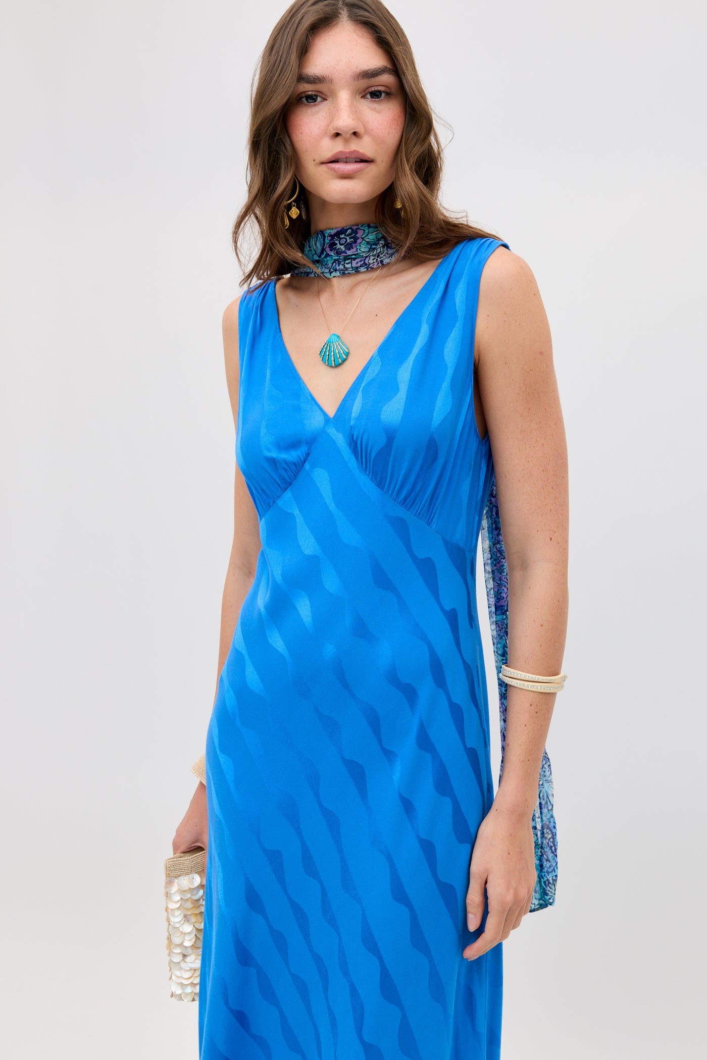A woman with wavy brown hair wears the sleeveless, V-neck Sandrine - Wave Jacquard Blue dress styled with a patterned scarf, white bangles, a shell necklace, and a beaded clutch.