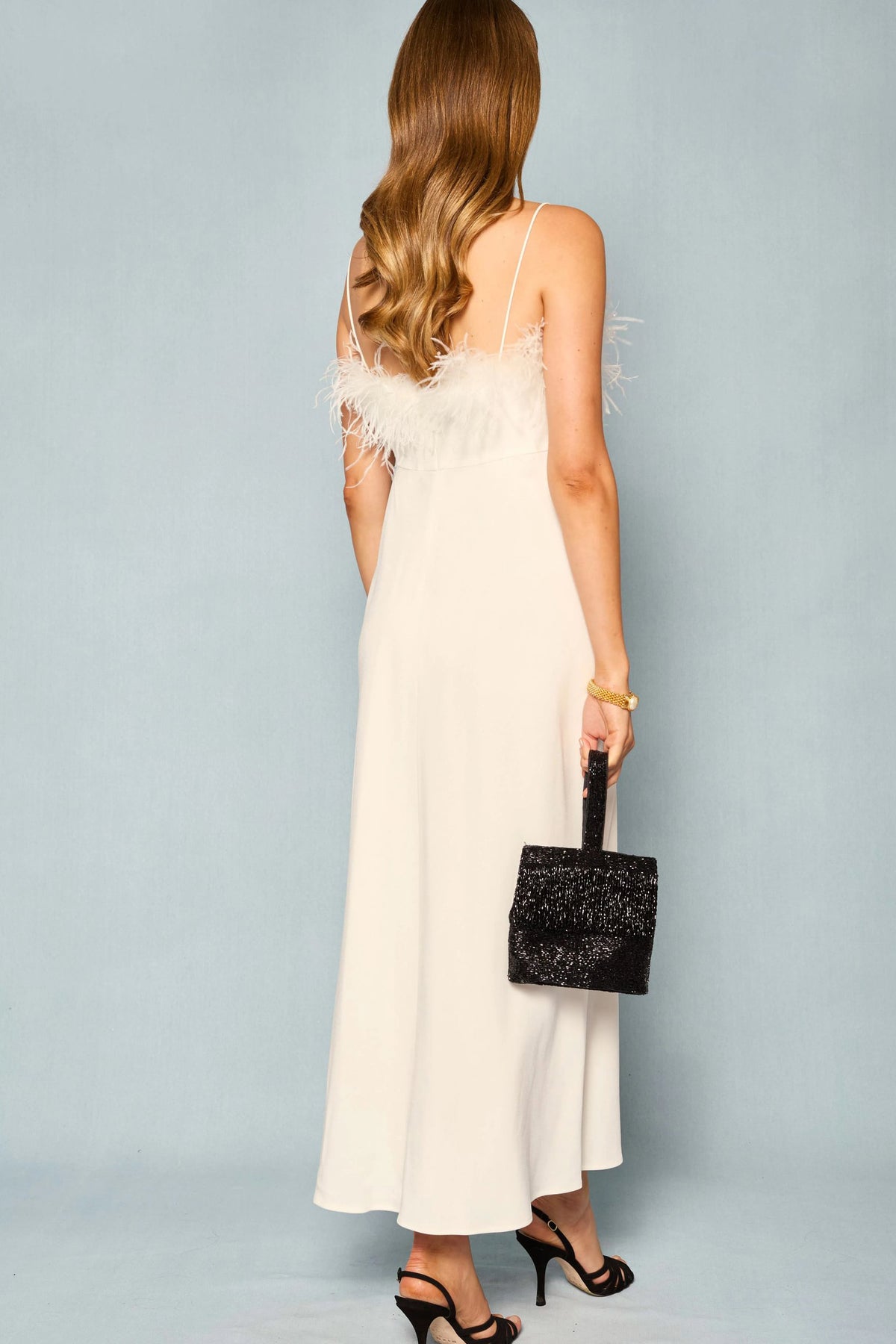 Woman with long wavy hair wearing the Selene - Ivory sleeveless dress with feather details, paired with black heeled sandals and a black textured handbag, standing against a light blue background.