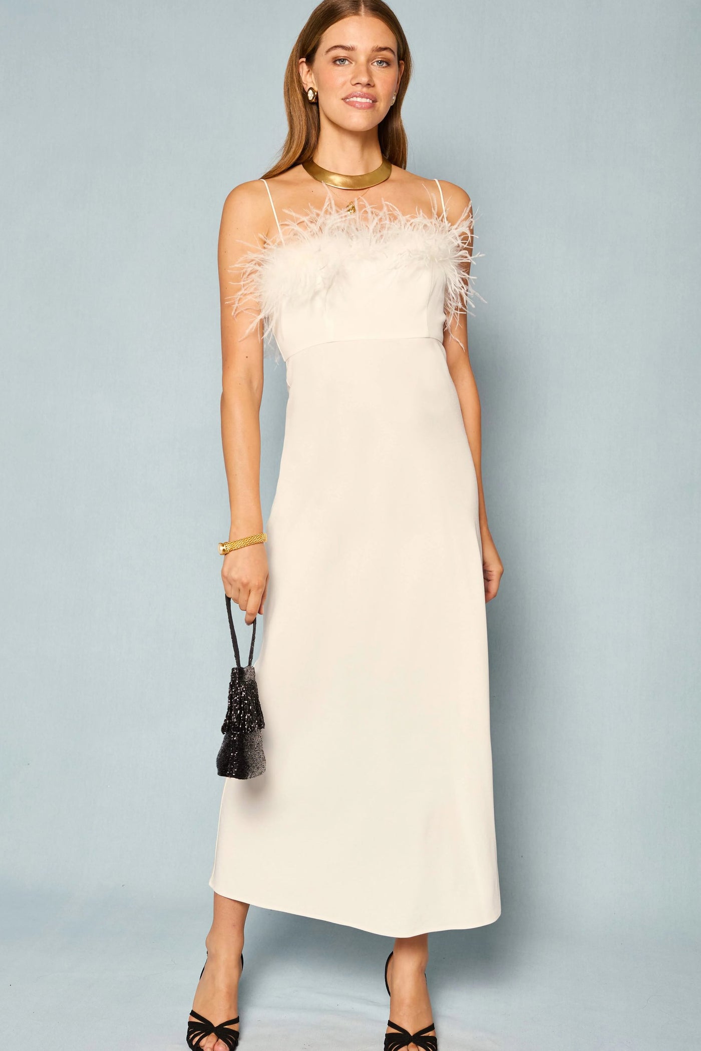 A woman stands against a plain light background wearing the Selene - Ivory sleeveless midi dress with feather detailing at the top, paired with black strappy heels and a small black purse.