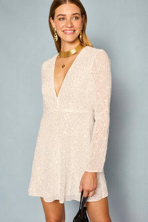 A woman is wearing the Sira - Squiggle Embellishment Ivory mini dress, which features long sleeves and a V-neckline with sparkling sequin details. She is smiling, holding a black bag, and accessorized with gold jewelry against a plain blue background.