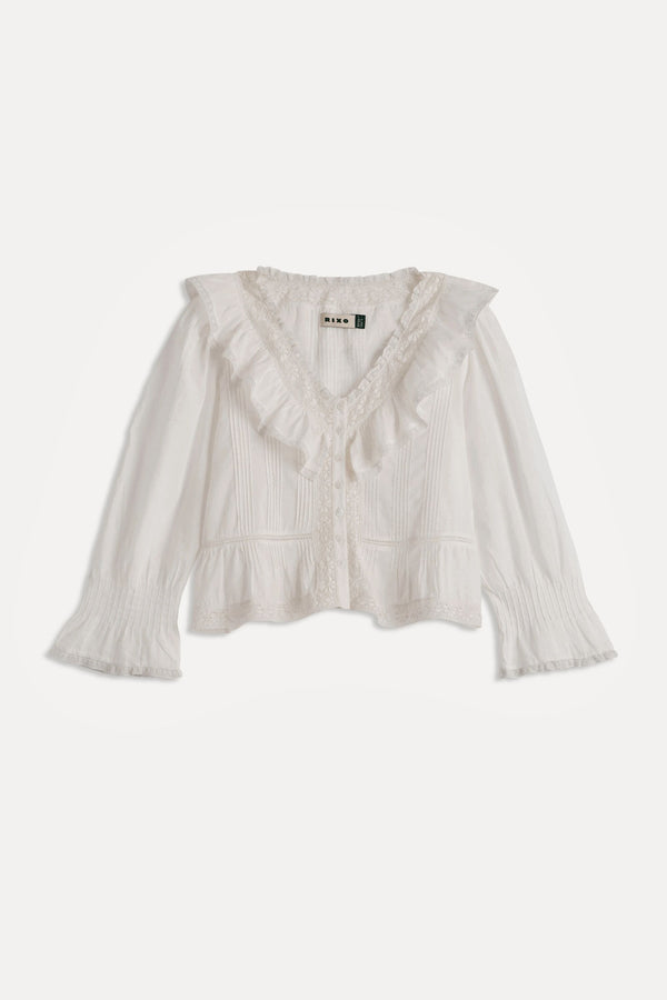 The Solene - Ivory is a white, long-sleeved blouse featuring a ruffled V-neckline, vintage-style pintuck detailing, flared textured cuffs, and a delicate feminine touch—perfect for fans of vintage-inspired fashion.