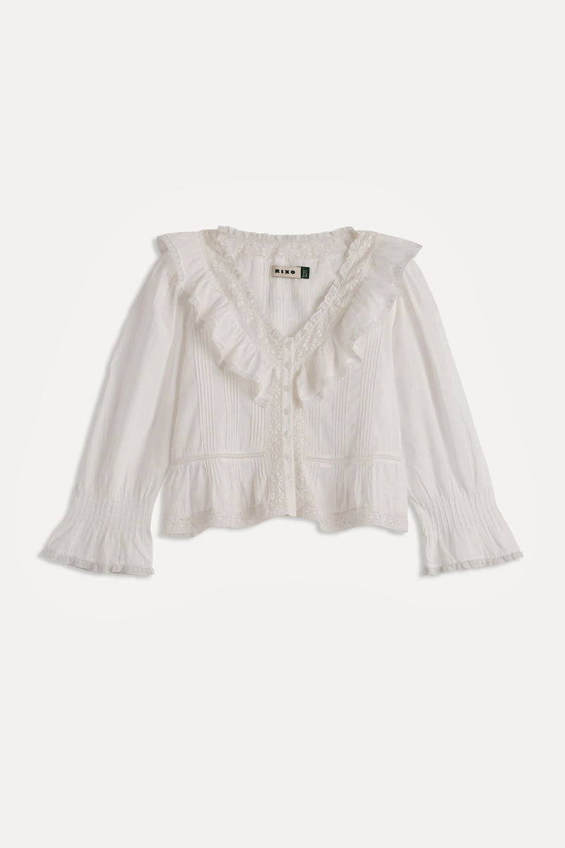 The Solene - Ivory is a white, long-sleeved blouse featuring a ruffled V-neckline, vintage-style pintuck detailing, flared textured cuffs, and a delicate feminine touch—perfect for fans of vintage-inspired fashion.