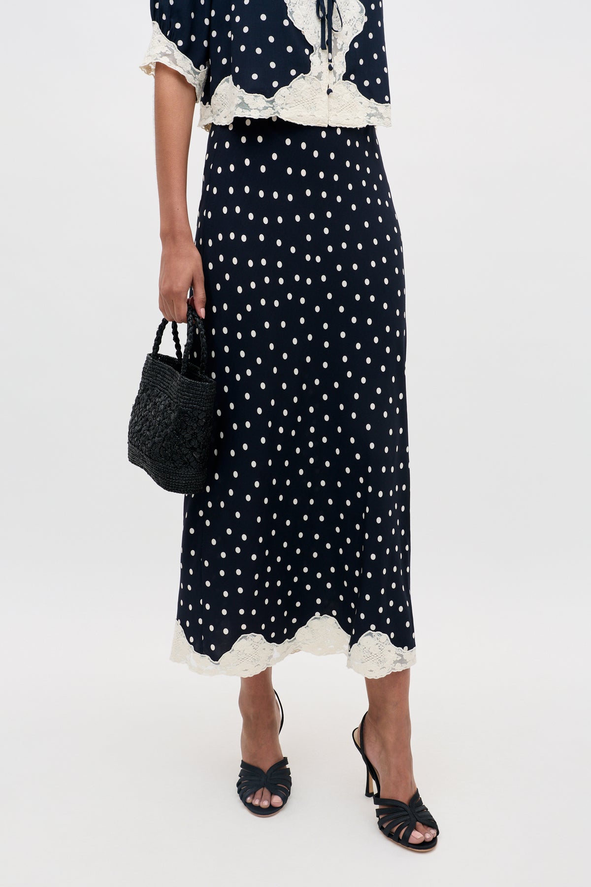 A woman wearing the Stassie - Layla Dot Navy skirt, paired with a matching top, black heeled sandals, and carrying a black woven handbag; only her lower half is visible against a plain background.