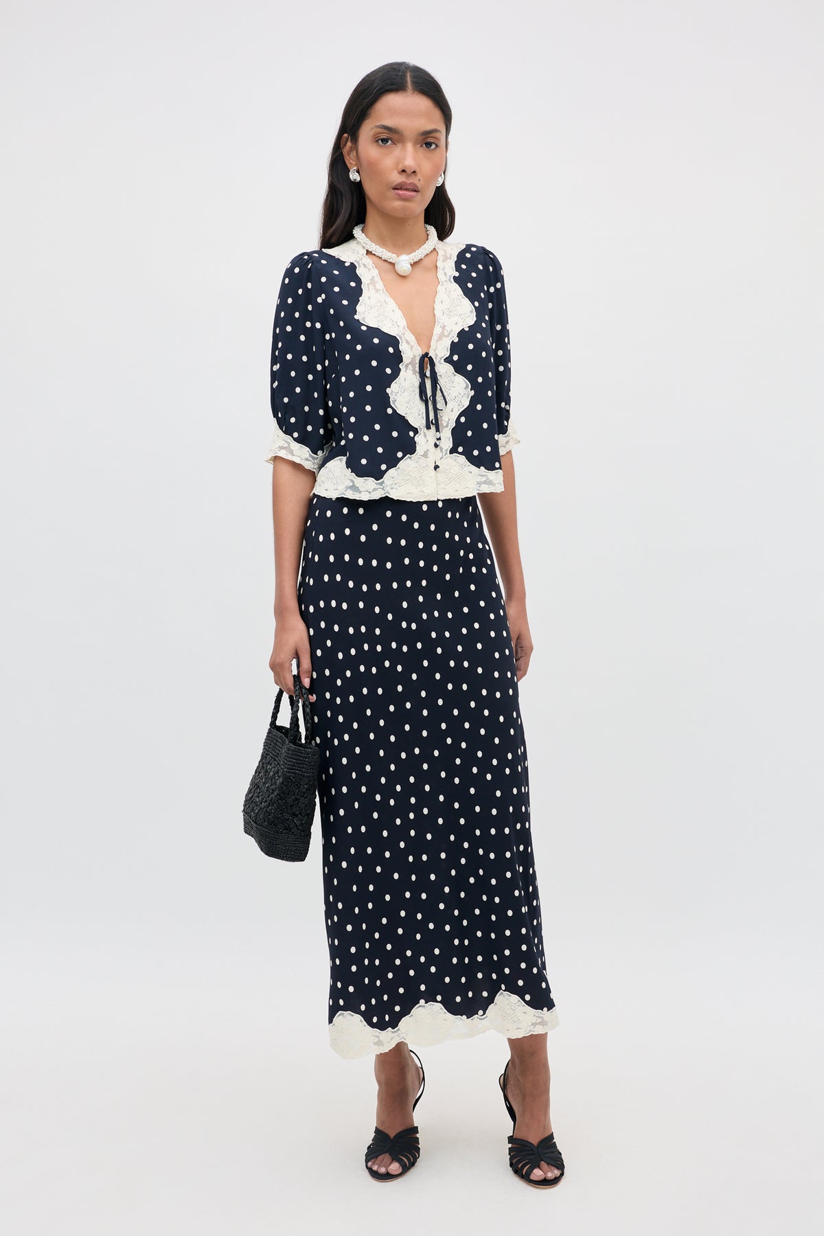 A woman with long dark hair and a neutral expression stands against a plain background wearing the Stassie - Layla Dot Navy two-piece outfit, which features navy blue polka dots and white lace trim. She completes her look with black sandals and a matching black handbag.