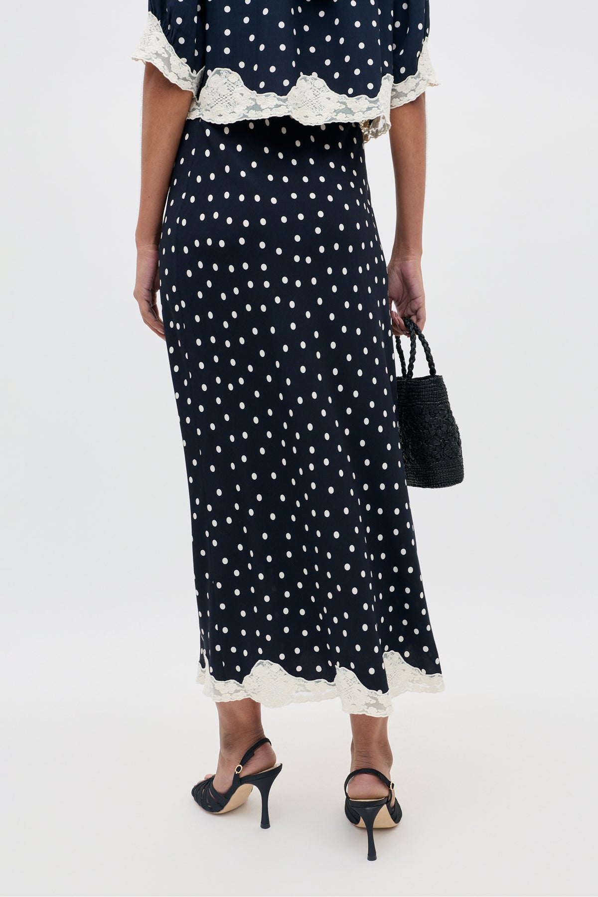 A woman stands facing away, wearing the Stassie - Layla Dot Navy skirt, which features white polka dots and cream lace trim. She pairs it with black heeled sandals and holds a small black woven handbag.