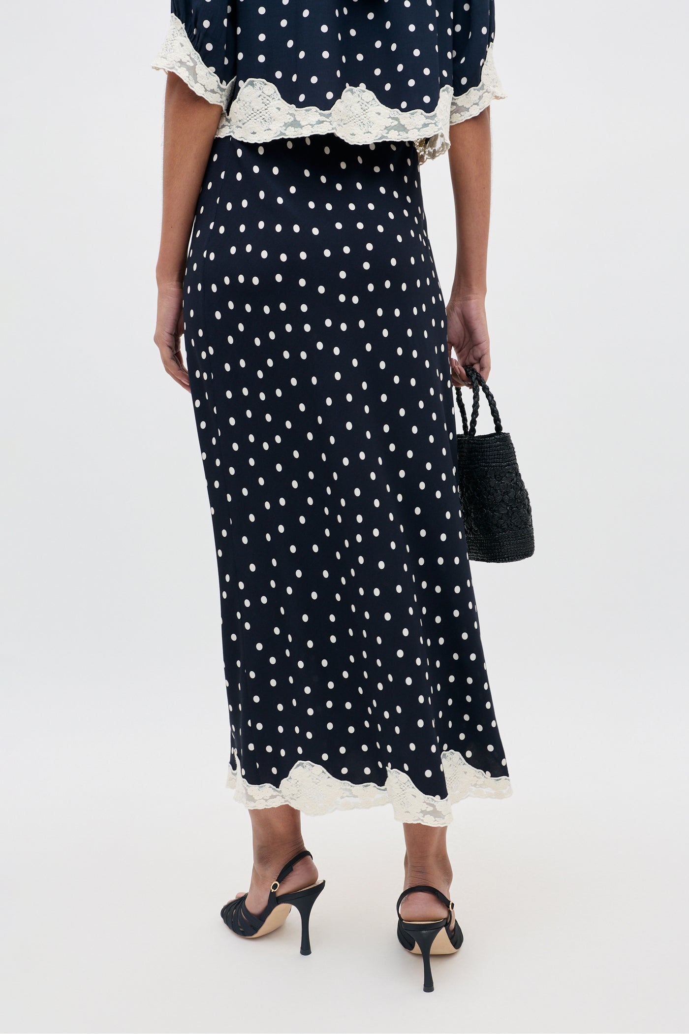 A woman stands facing away, wearing the Stassie - Layla Dot Navy skirt, which features white polka dots and cream lace trim. She pairs it with black heeled sandals and holds a small black woven handbag.
