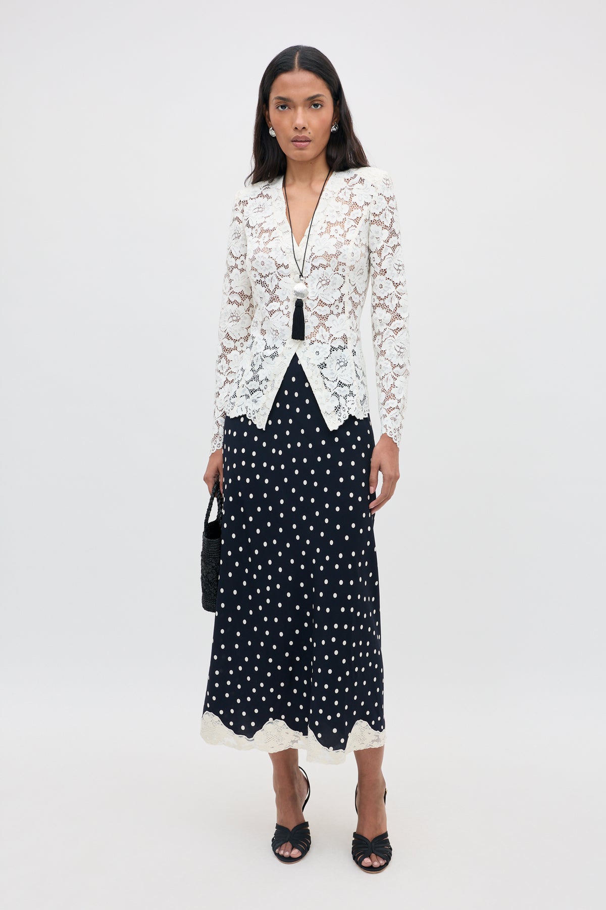 A woman stands against a plain background, wearing the Stassie - Layla Dot Navy midi skirt with black and white polka dots, paired with a white lace blazer, black sandals, and a long necklace. She holds a black handbag and has a neutral expression.