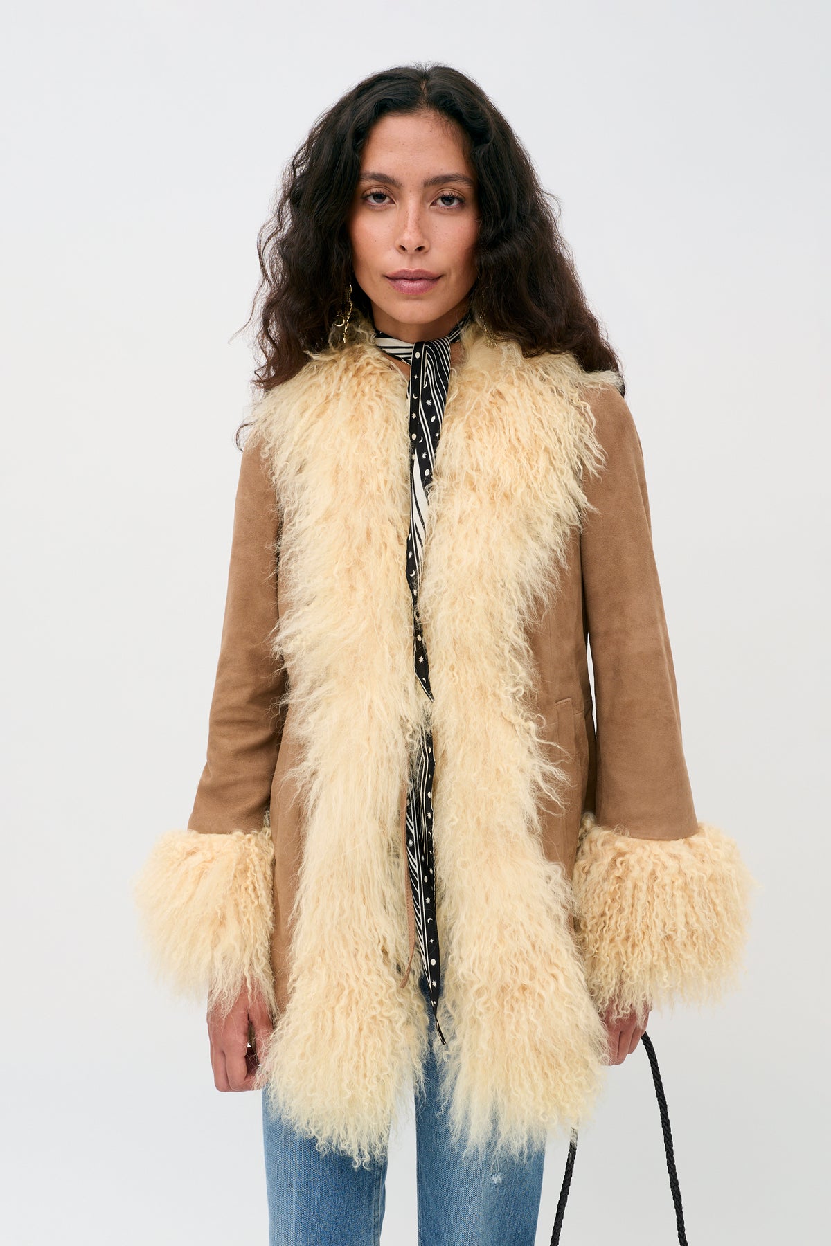 A woman with long curly hair wears the Nellie - Tan suede coat, featuring cream-colored shaggy fur trim on the collar, front, and cuffs, over a patterned shirt and blue jeans, standing against a plain light background.