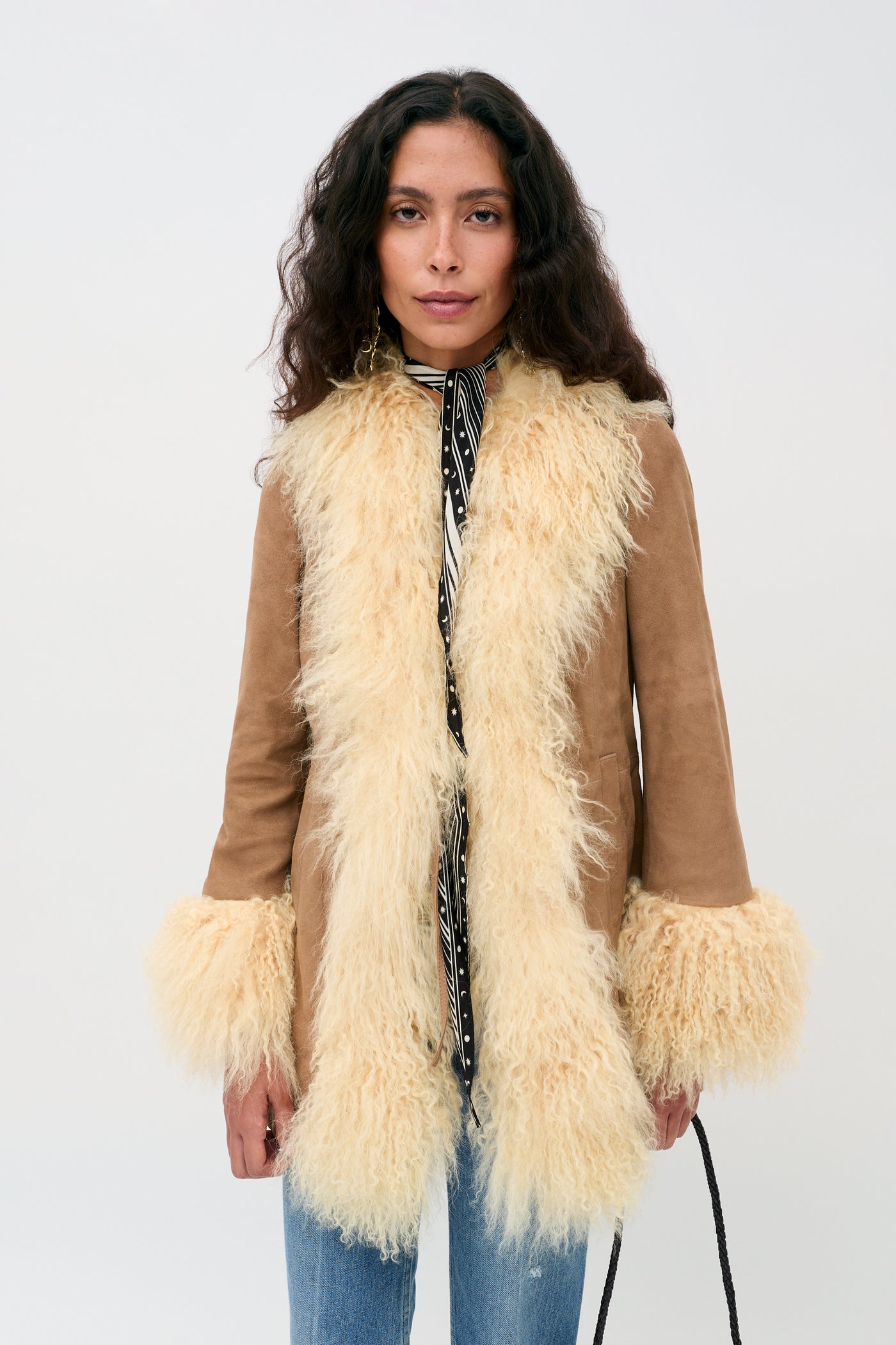 A woman with long curly hair wears the Nellie - Tan suede coat, featuring cream-colored shaggy fur trim on the collar, front, and cuffs, over a patterned shirt and blue jeans, standing against a plain light background.