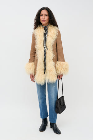 Wearing the Nellie - Tan suede coat with fluffy cream trim, blue jeans, black ankle boots, a black patterned scarf, and carrying a black woven bag, a person stands against a plain white background.