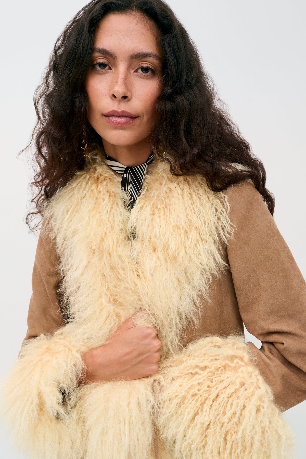 A woman with long curly hair wears the Nellie - Tan suede coat featuring cream-colored shearling trim and a black-and-white striped scarf, posing against a plain light background.