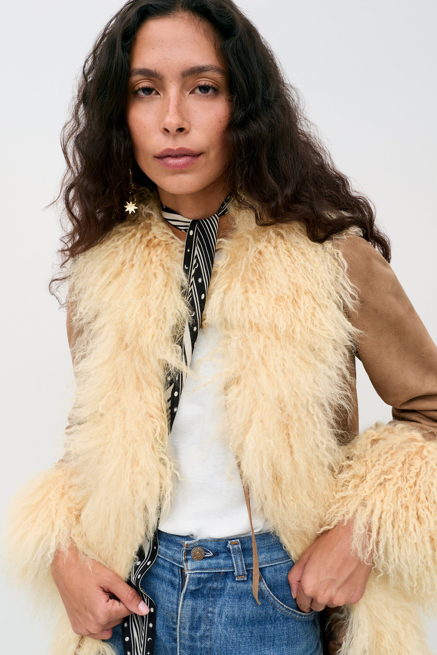 A person with long wavy hair wears the Nellie - Tan shearling jacket, a white shirt, blue jeans, a striped scarf, and a star-shaped earring, standing against a plain light background.