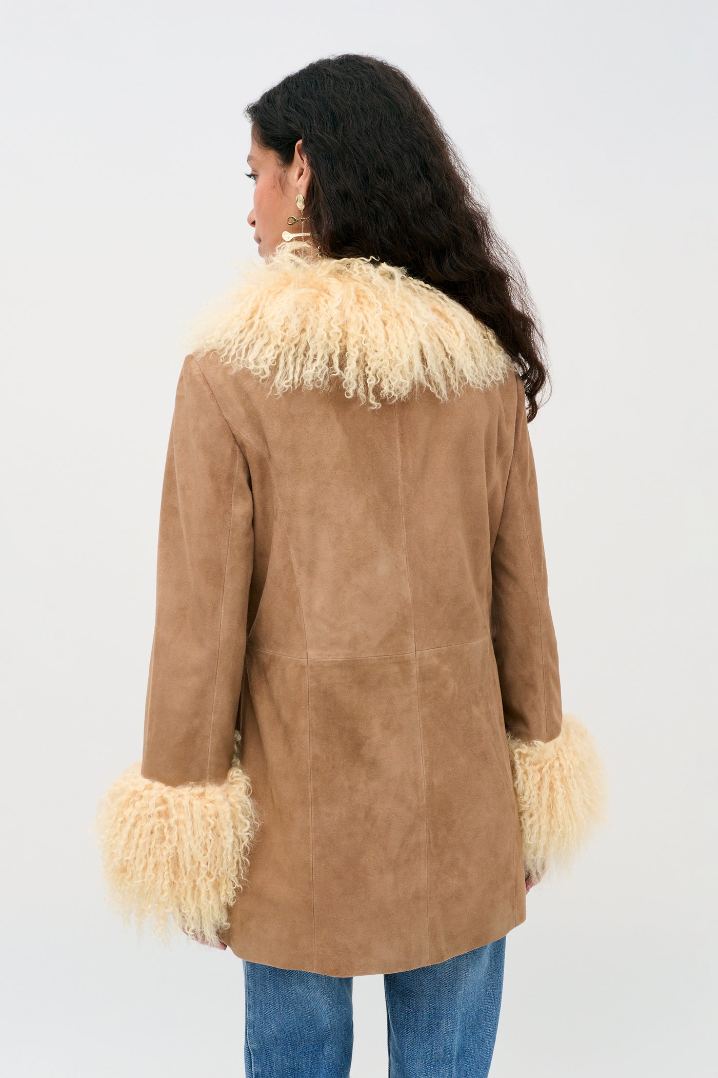 A woman with long, dark wavy hair is seen from the back, modeling the Nellie - Tan jacket—a tan suede piece featuring cream-colored, fluffy shearling at the collar and cuffs. She pairs it with blue jeans and stands against a plain light background.