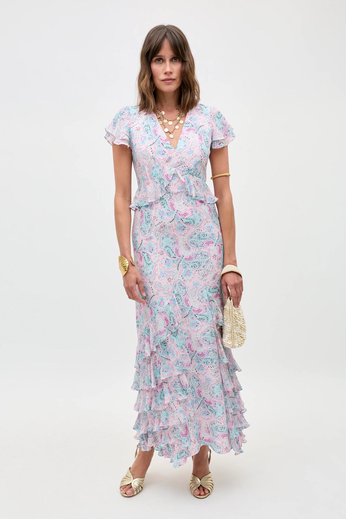 A woman stands against a plain background wearing the Thanvi - Butterfly Paisley Cream dress—a short-sleeve, V-neck ruffled maxi—paired with gold sandals, layered necklaces, a gold bracelet, and a small textured handbag.
