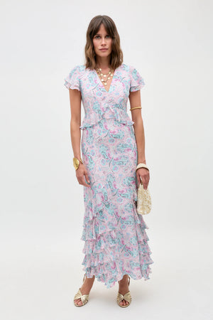 A woman stands against a plain background wearing the Thanvi - Butterfly Paisley Cream dress—a short-sleeve, V-neck ruffled maxi—paired with gold sandals, layered necklaces, a gold bracelet, and a small textured handbag.