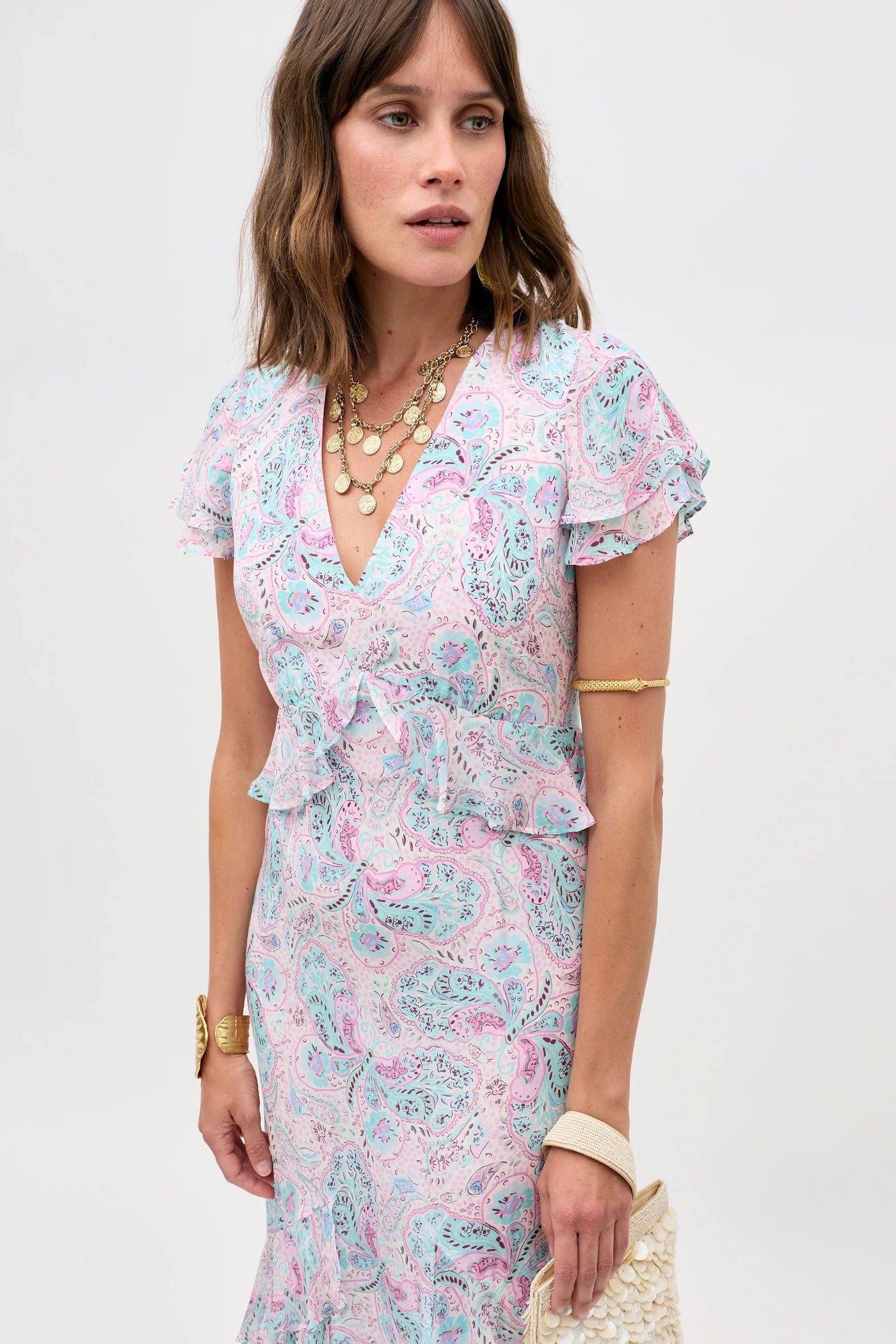 Wearing the Thanvi - Butterfly Paisley Cream dress with short ruffled sleeves, she accessorizes with layered gold necklaces, bracelets, and a textured clutch—creating an ideal summer party look against a simple light background.
