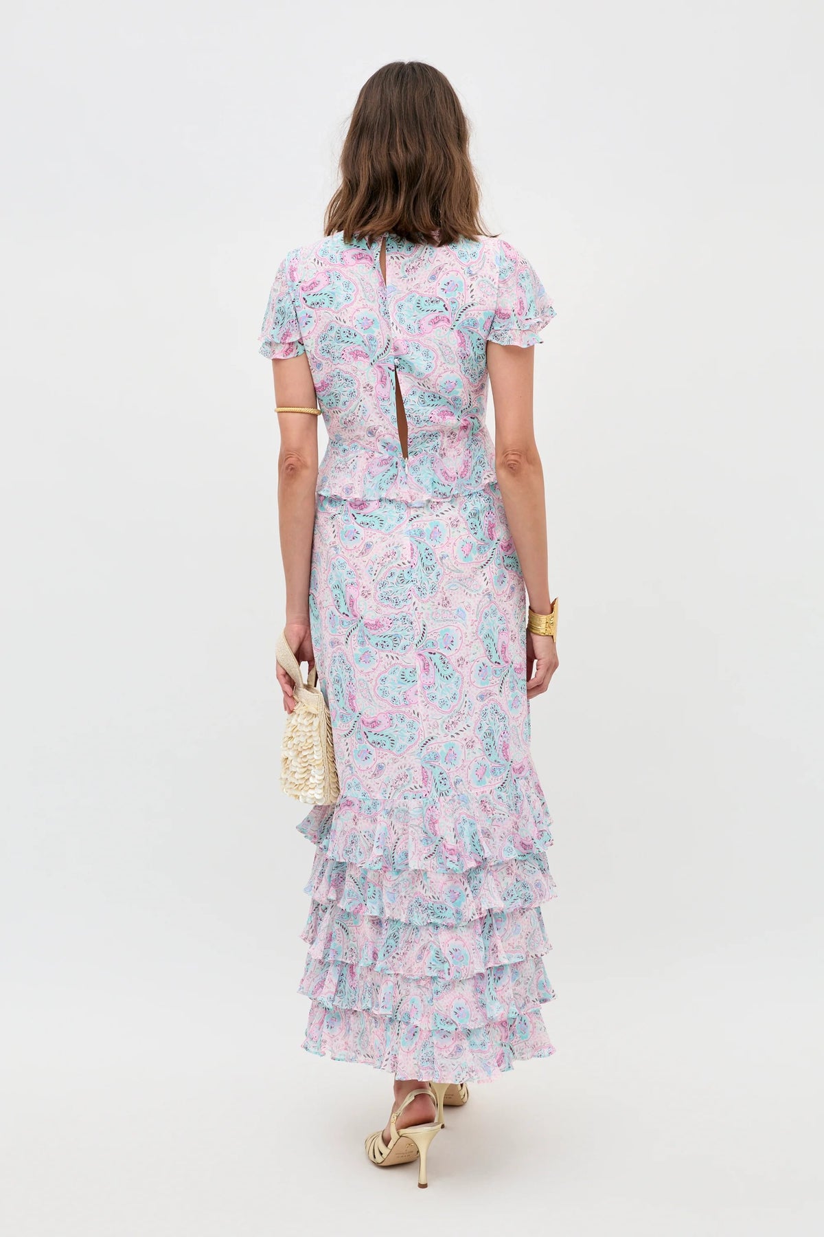 A woman faces away, wearing the Thanvi - Butterfly Paisley Cream dress with ruffled hem tiers. She holds a cream woven clutch, accessorized with gold heels and jewelry. The background is plain and light-colored.