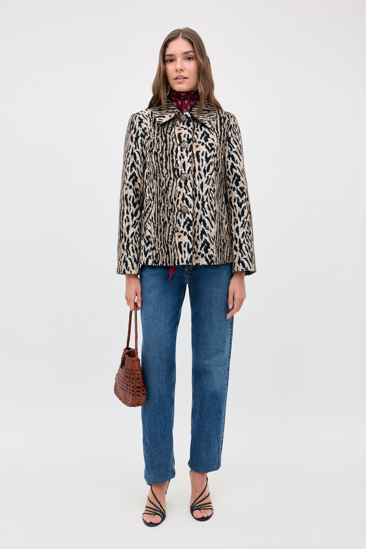 A woman stands facing forward against a plain background, wearing the Tommie - Bohemia Leopard jacket, paired with blue jeans, black strappy heels, and holding a brown woven handbag. She has straight brown hair and a neutral expression.