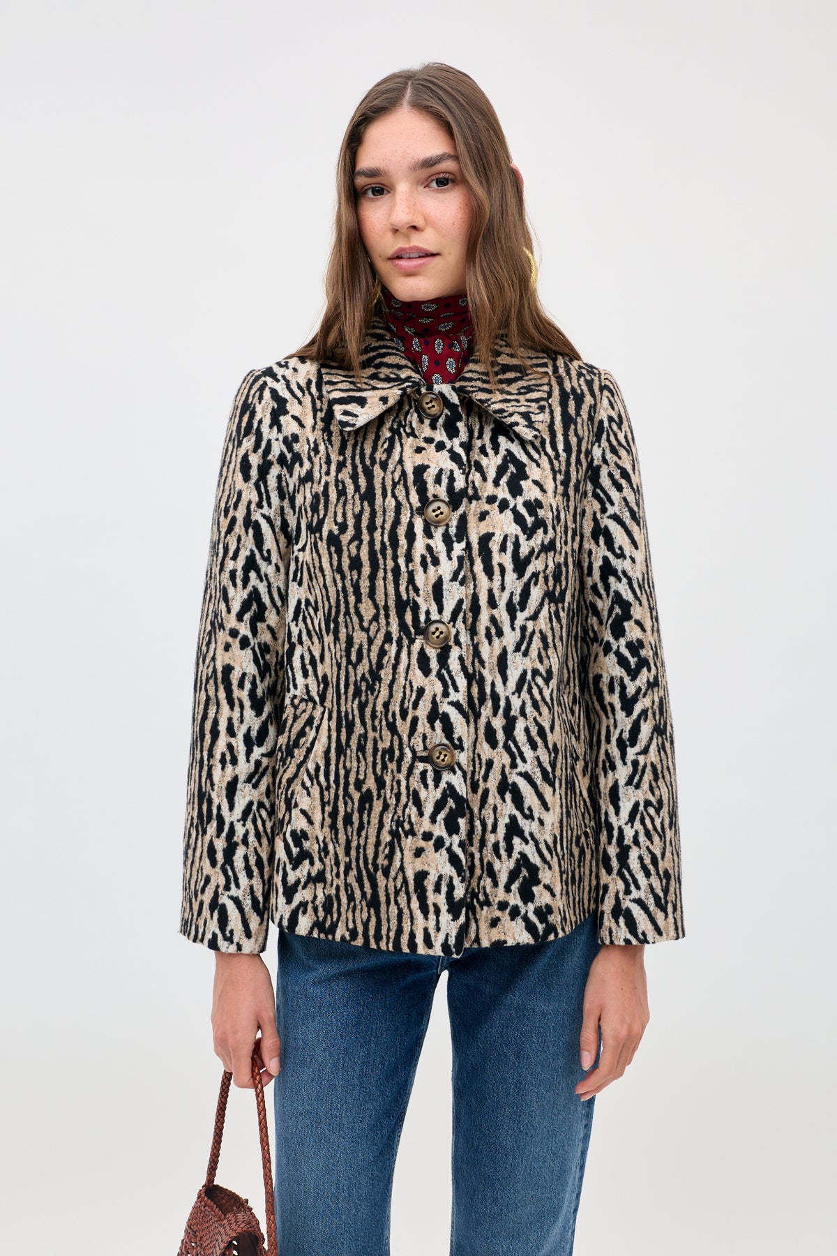 A woman wearing the Tommie - Bohemia Leopard jacket with blue jeans and a red patterned scarf holds a small brown bag against a plain light background.