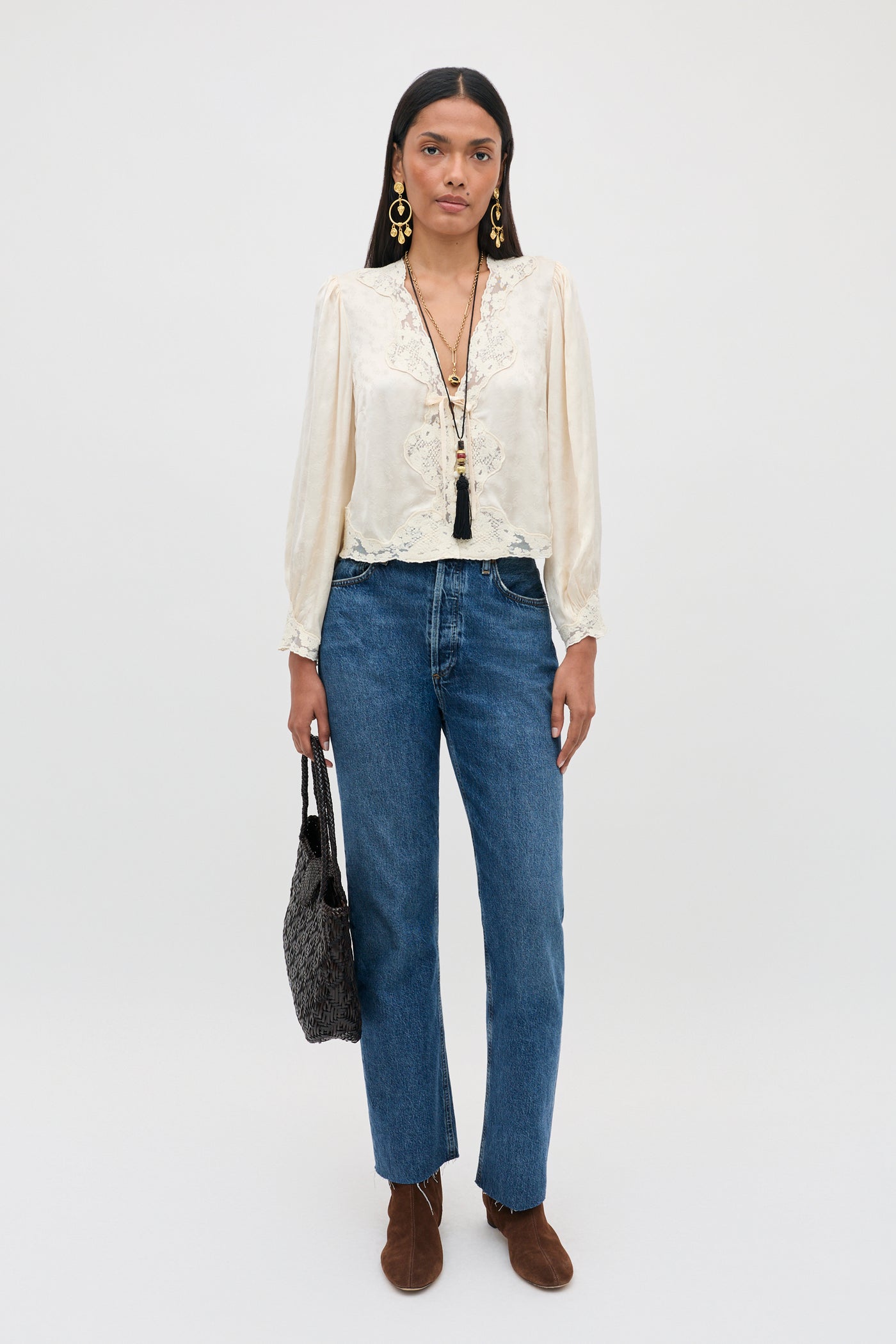 A woman stands against a plain background, wearing the Tove - Floral Trail Jacquard Champagne blouse paired with blue jeans, brown boots, statement earrings, layered necklaces, and holding a textured black bag.
