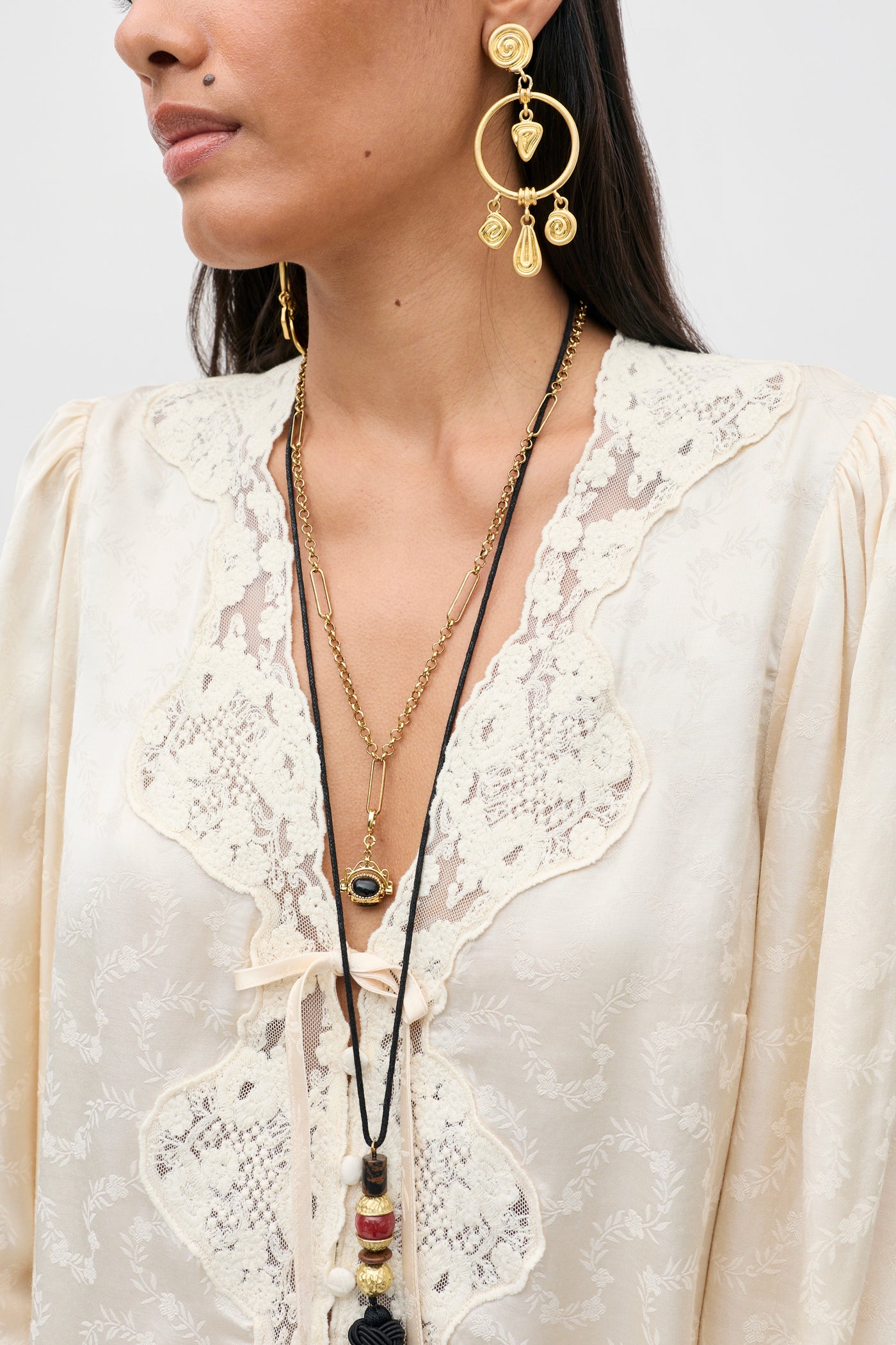 A woman wears the Tove - Floral Trail Jacquard Champagne dress, styled with layered gold necklaces and large, ornate gold earrings. The image is cropped from her shoulders to mid-chest against a neutral background.