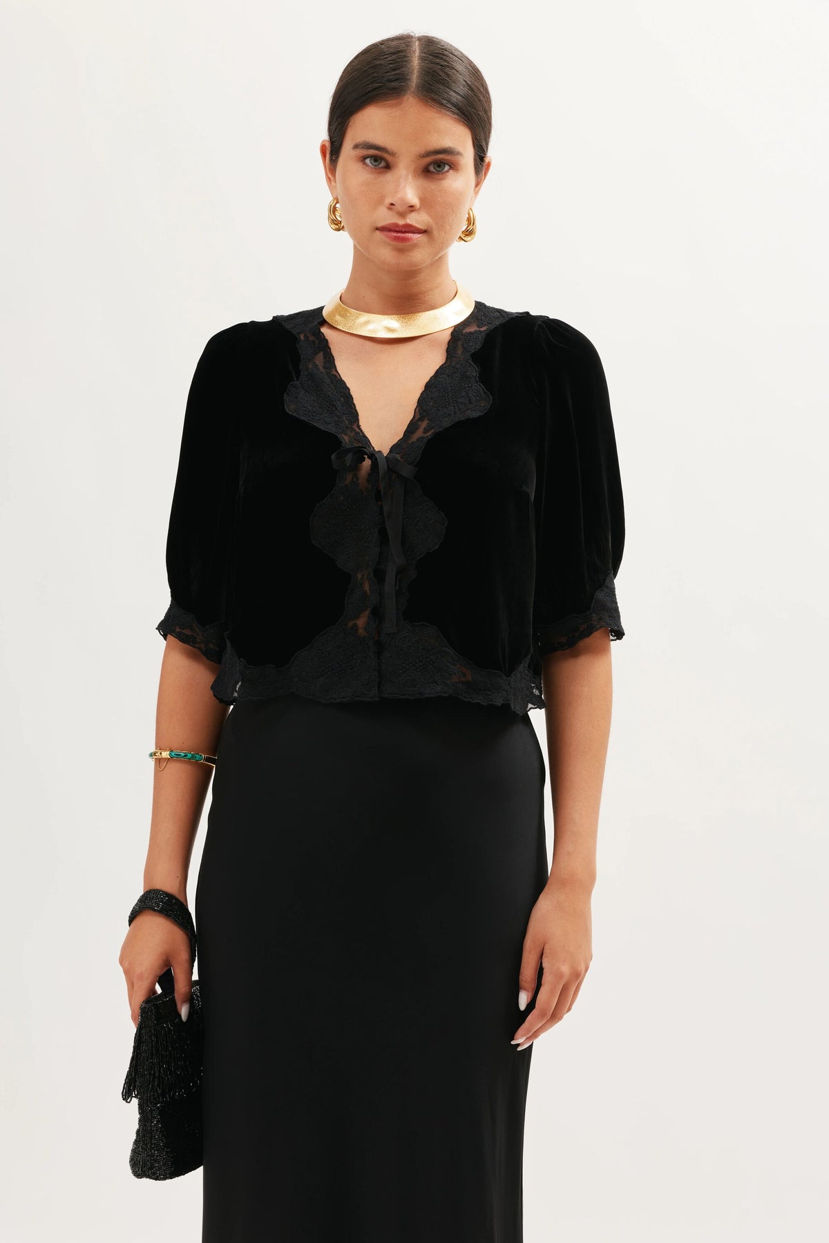 A woman wears the Ursula - Black velvet blouse with lace and ruffle trim, short sleeves, and a long black skirt. She accessorizes with gold jewelry and carries a black bag.