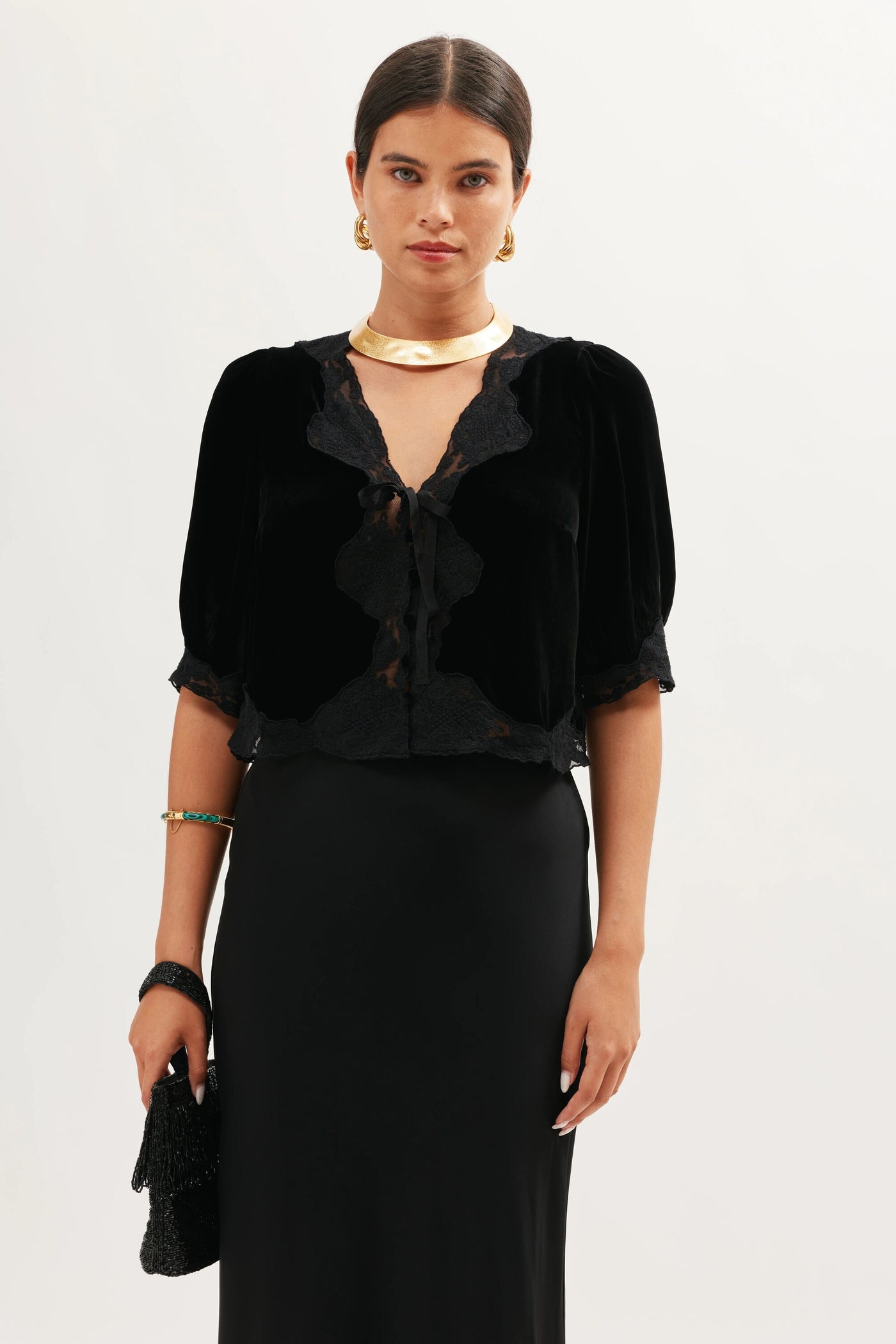 A woman wears the Ursula - Black velvet blouse with lace and ruffle trim, short sleeves, and a long black skirt. She accessorizes with gold jewelry and carries a black bag.