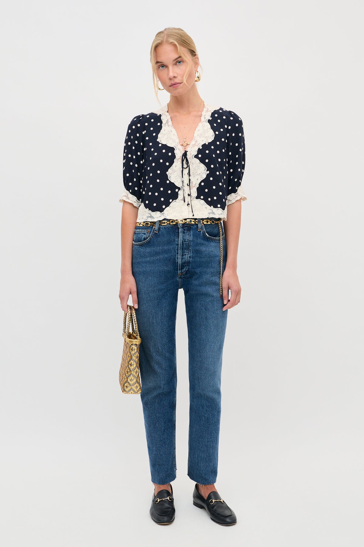 A woman stands against a plain background wearing the ursula top layla dot navy—a navy polka-dot blouse with lace trim—paired with blue jeans, black loafers, and holding a gold patterned handbag. She has blonde hair tied back and is wearing gold earrings.