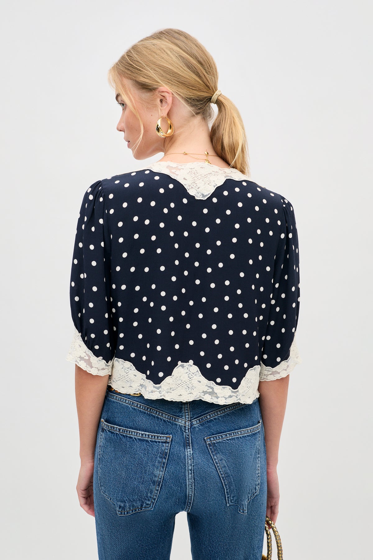 A woman with blonde hair in a ponytail is seen from the back wearing the ursula top layla dot navy with blue jeans and holding a small handbag.
