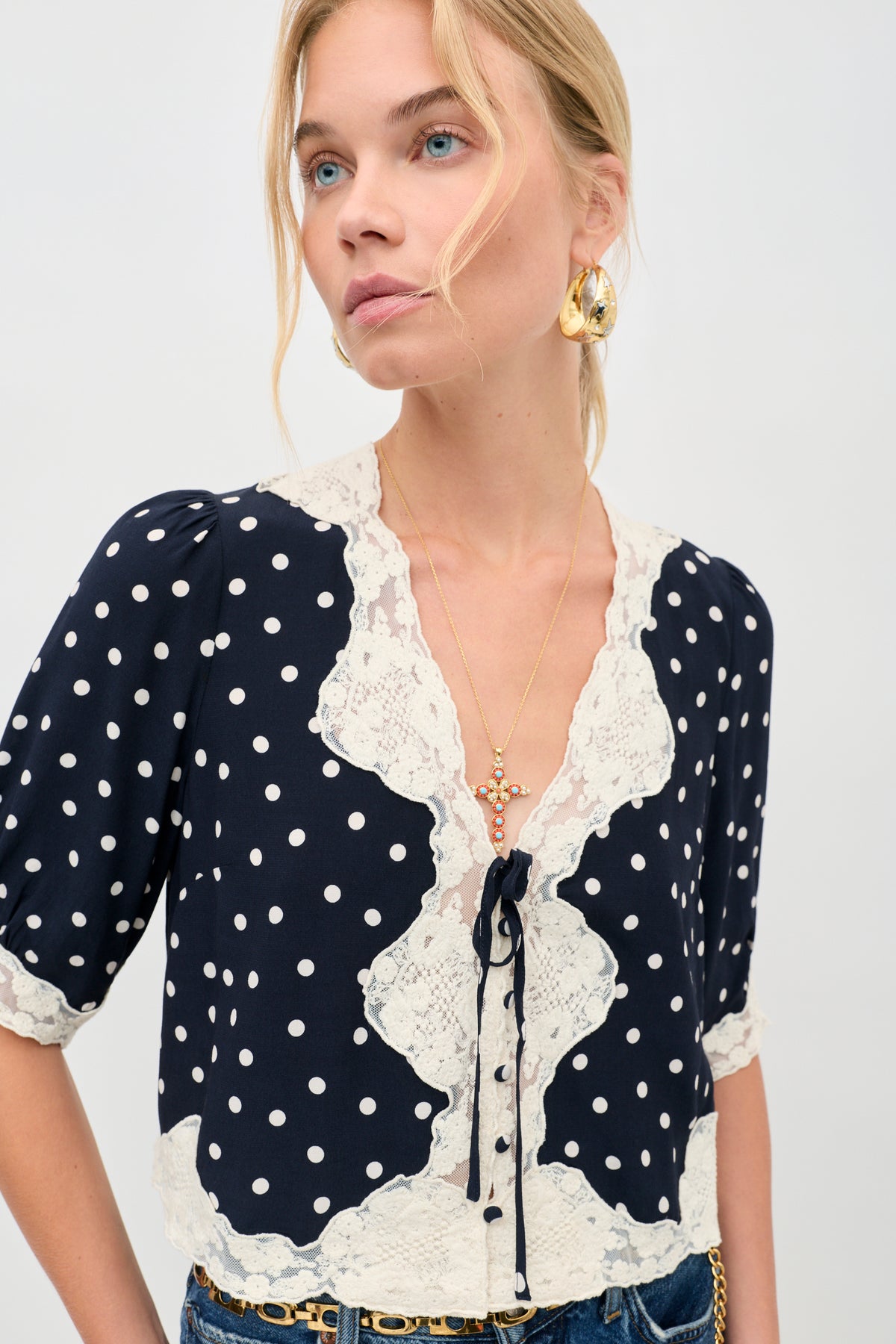 A woman wears the “ursula top layla dot navy,” a navy blouse with white polka dots and cream lace trim, styled with gold earrings and a necklace. Her blonde hair falls loosely as she gazes to the side against a plain backdrop.