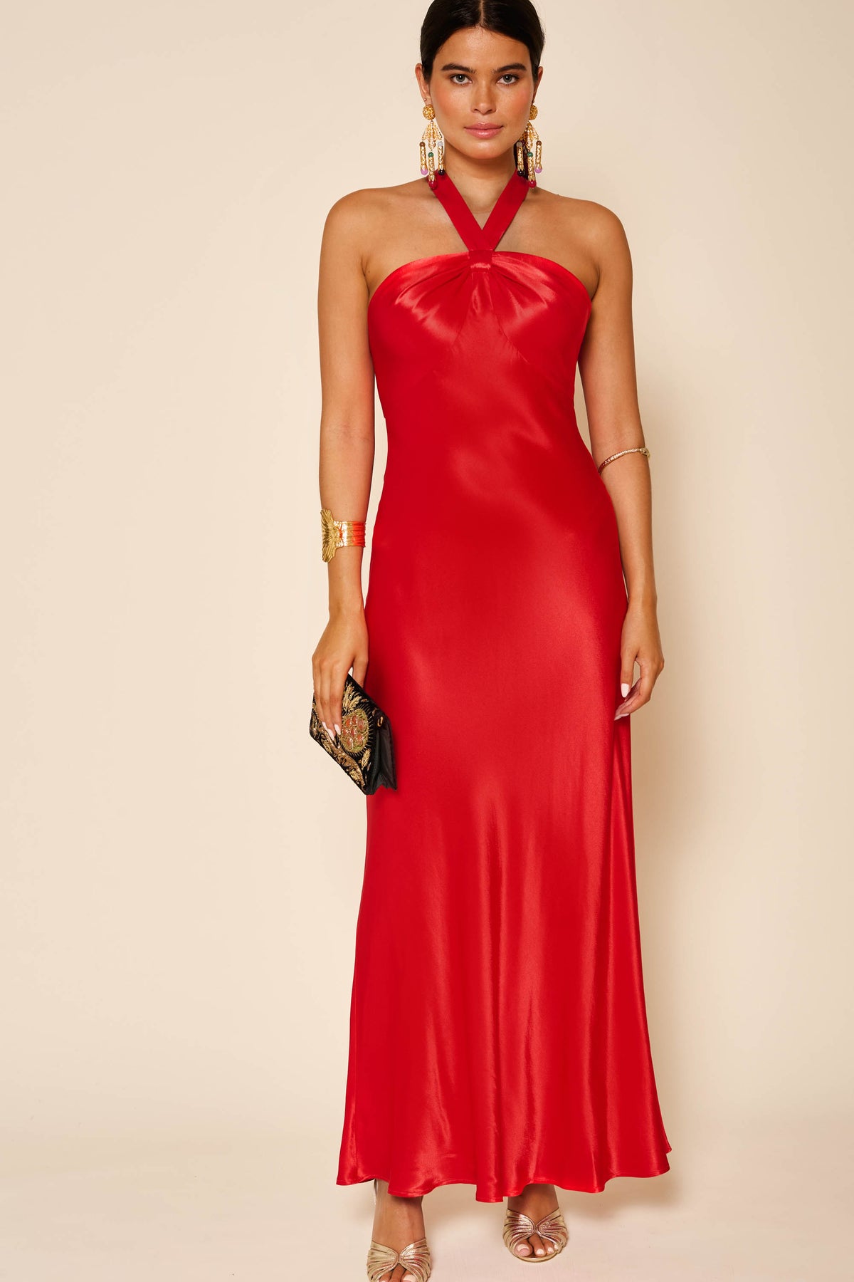 A woman wears the Losanna - Red long, open back satin dress with gold earrings, bracelets, rings, and holds a black clutch against a neutral background.