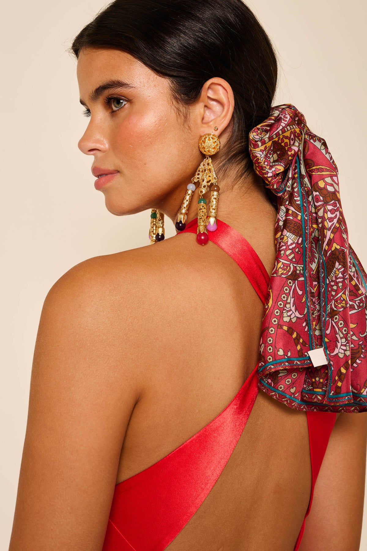 A woman with dark hair in a low ponytail with a colorful scarf poses against a neutral background, wearing the Losanna - Red satin open-back dress and large dangling earrings.