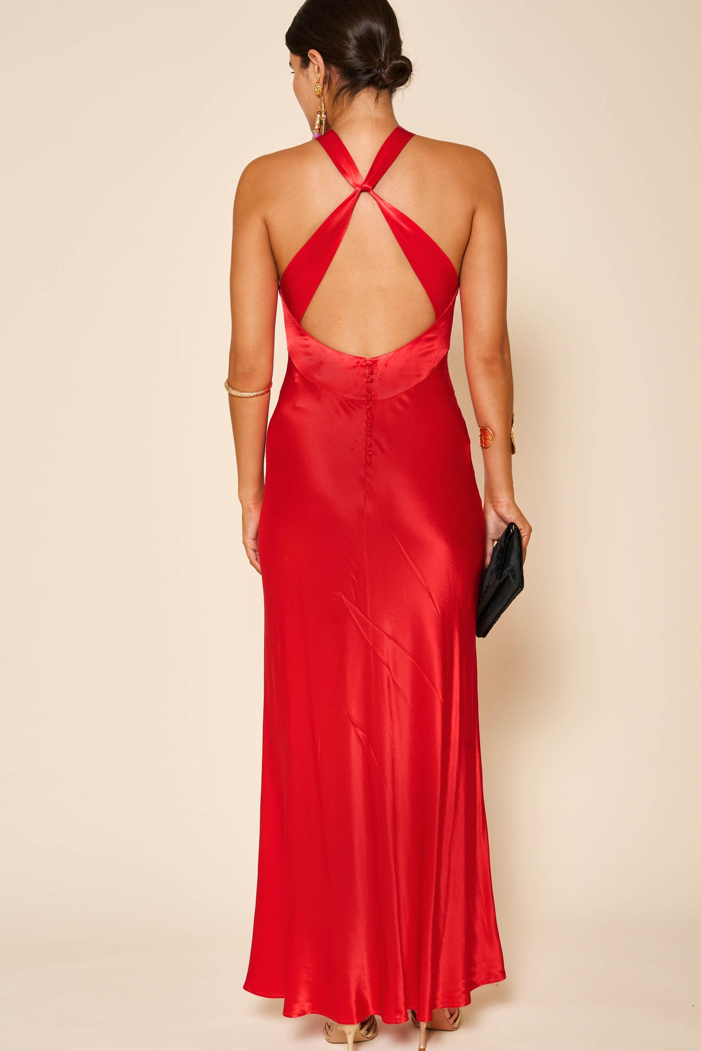 A woman faces away in the Losanna - Red dress, featuring satin fabric, a criss-cross halter, and open back. She holds a black clutch, wears gold jewelry, and her hair is styled in a low bun.