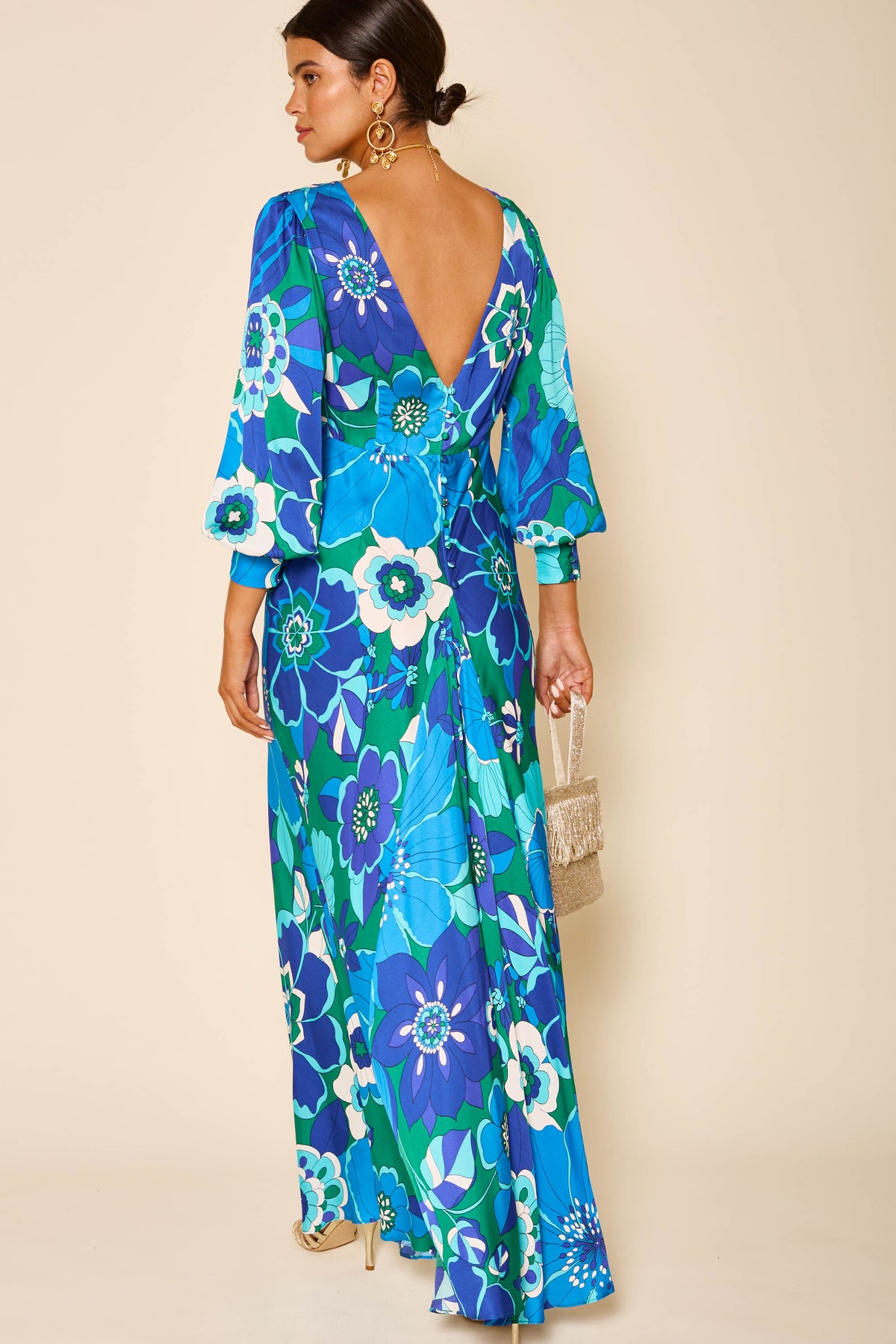 A woman in the Marni Miami Floral Emerald gown with blouson sleeves and a deep V-back stands facing away, holding a small woven handbag. Gold earrings catch the light and her hair is styled in a low bun.