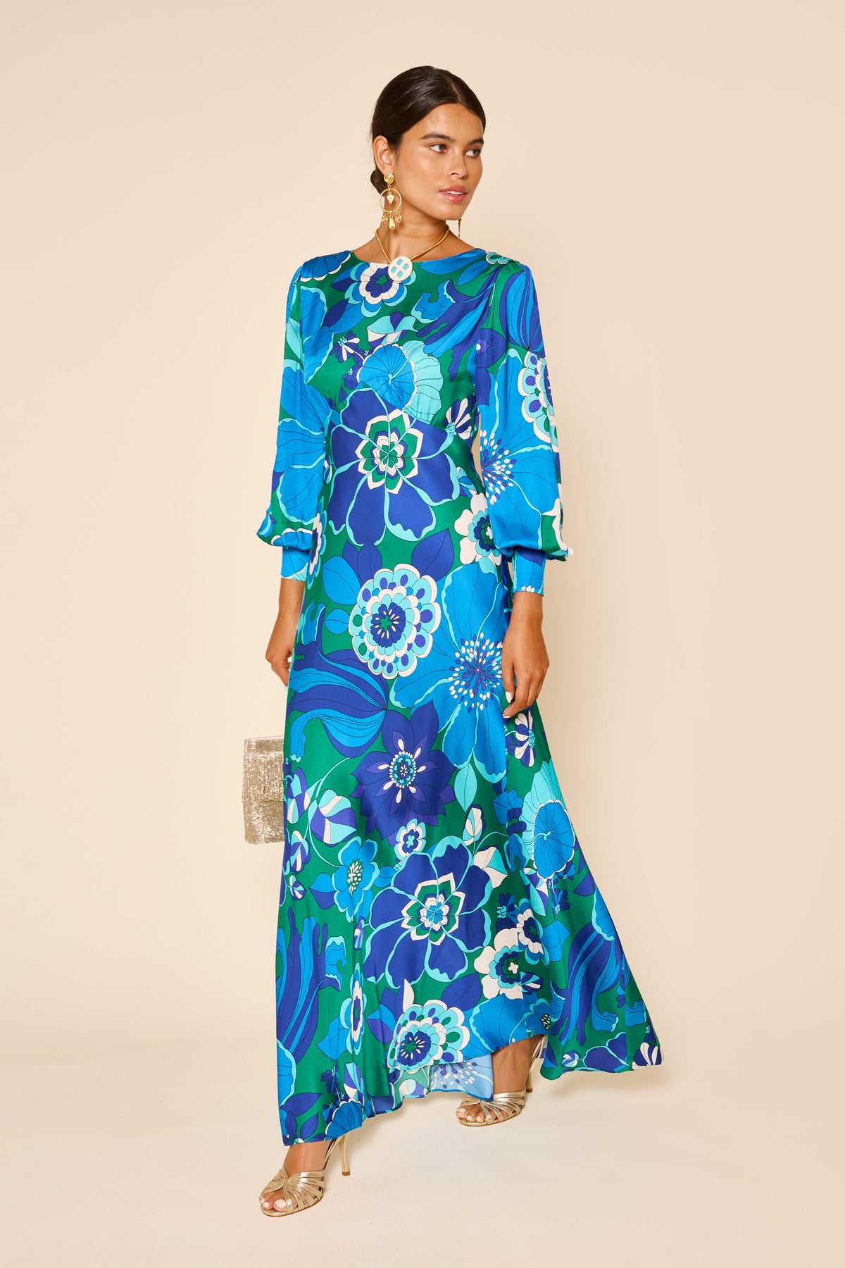 A woman in the marni - miami floral emerald gown, featuring blouson sleeves, gold earrings and sandals, holds a small clutch while standing against a plain beige background.
