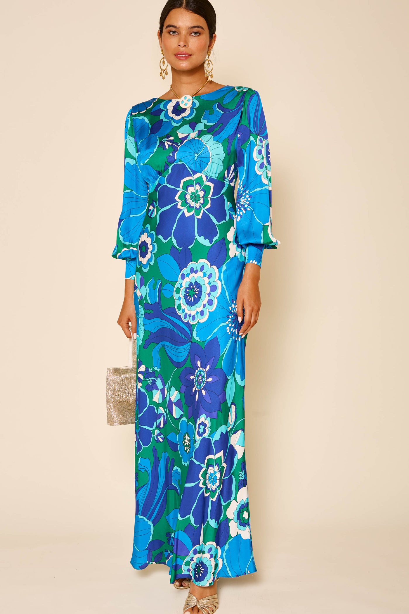A woman in the marni - miami floral emerald gown with blouson sleeves in blue and green holds a glittery clutch, wears gold earrings, has her hair tied back, and stands against a light beige background.
